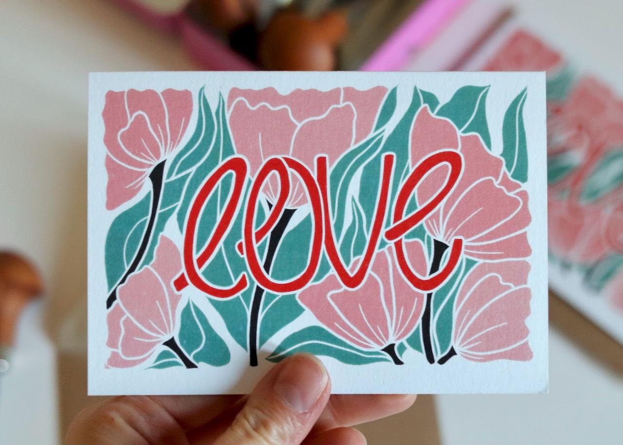 A colourful floral card featuring pink modern flowers, flowing green leaves with the word "love" in a warm red colour. An original design created using linocut.