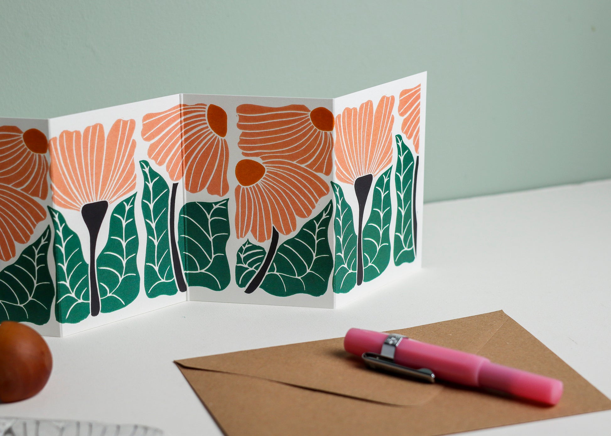 Garden Chorus Garland card with C6 kraft envelope. Hand-carved botanical portrait greeting card in peach and green on recycled cardstock, photographed with eco-friendly envelope, ready for posting.
