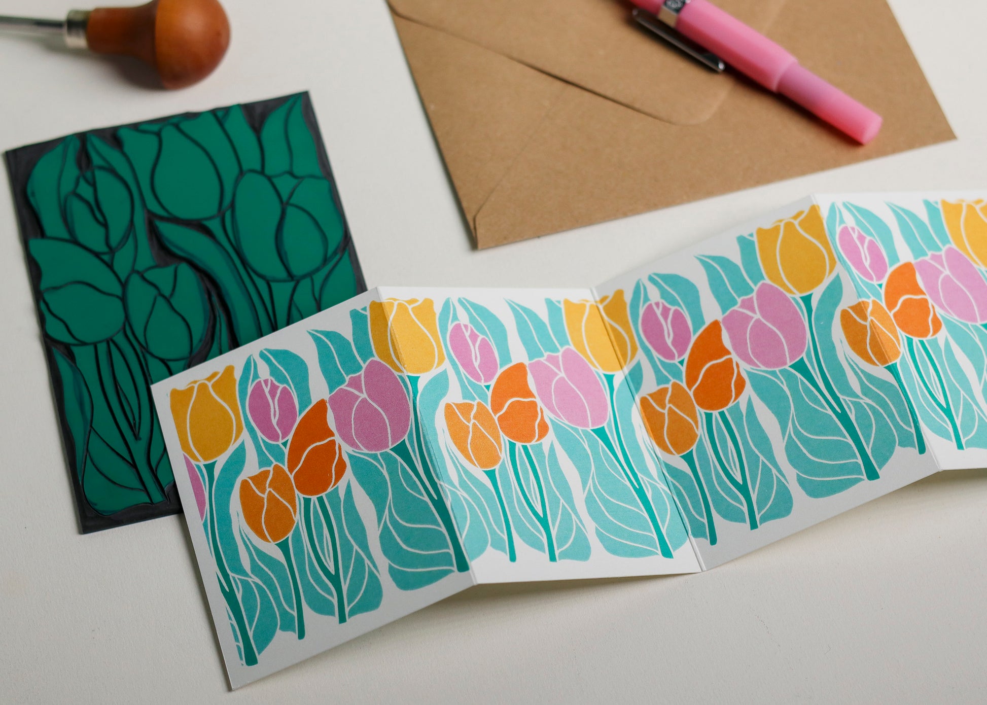 Hand-carved lino block shown beside finished Spring Tulip Garland card. Traditional linocut printmaking tools and original carved linoleum block displayed with the botanical portrait card. No AI, authentic craftsmanship from Norfolk studio.