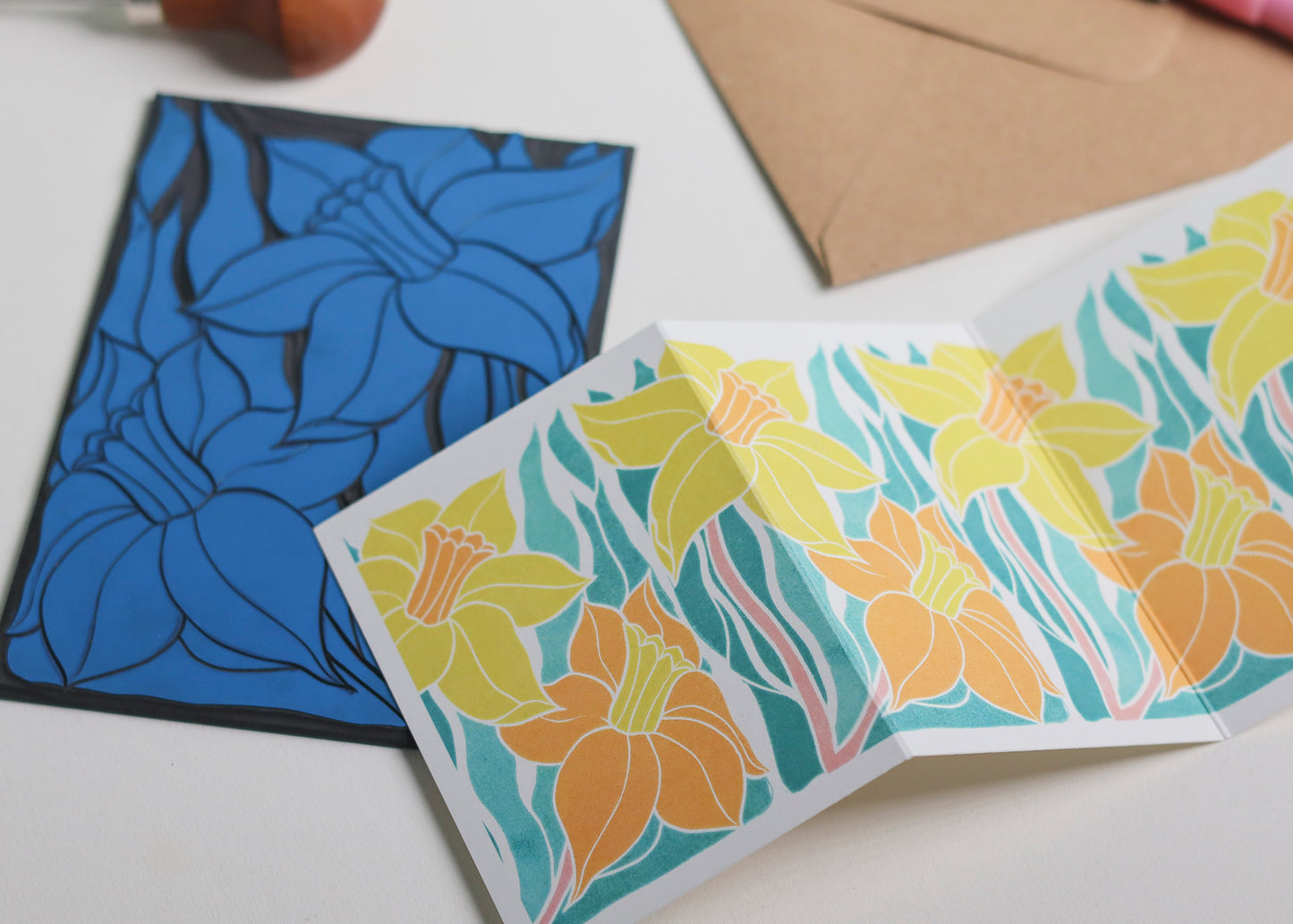 Hand-carved lino block shown beside finished Welsh Daffodil Garland card. Traditional linocut printmaking tools and original carved linoleum block displayed with contemporary yellow and green  botanical portrait card. No AI, authentic craftsmanship from Norfolk studio