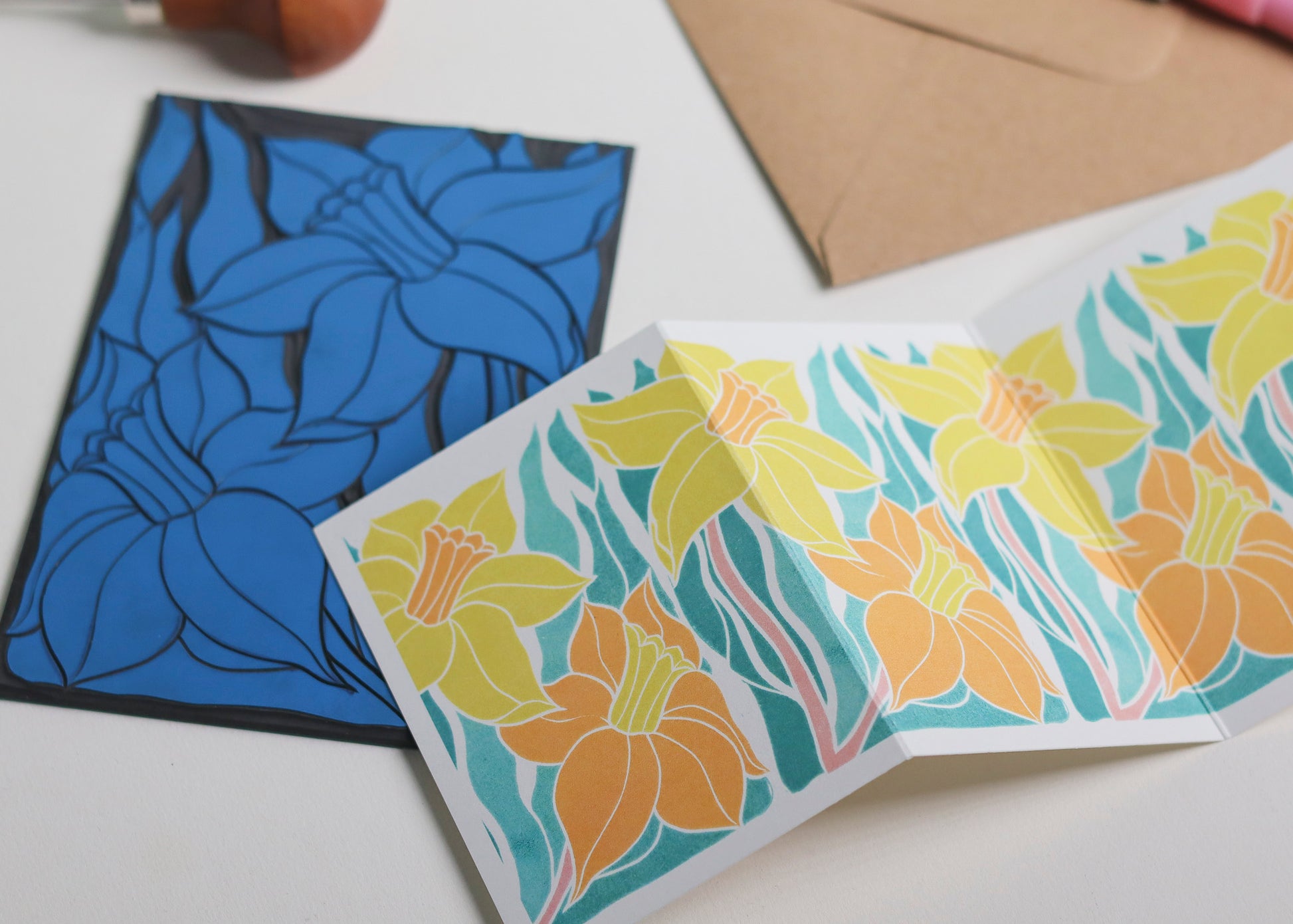 Hand-carved lino block shown beside finished Welsh Daffodil Garland card. Traditional linocut printmaking tools and original carved linoleum block displayed with contemporary yellow and green  botanical portrait card. No AI, authentic craftsmanship from Norfolk studio