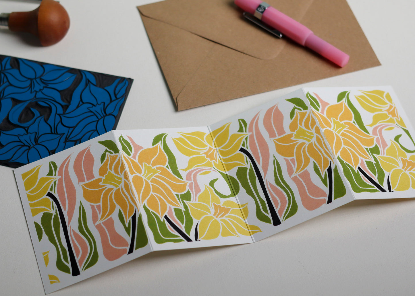 Hand-carved lino block shown beside finished Daffodil Garland card. Traditional linocut printmaking tools and original carved linoleum block displayed with a contemporary botanical portrait card. No AI, authentic craftsmanship from Norfolk studio