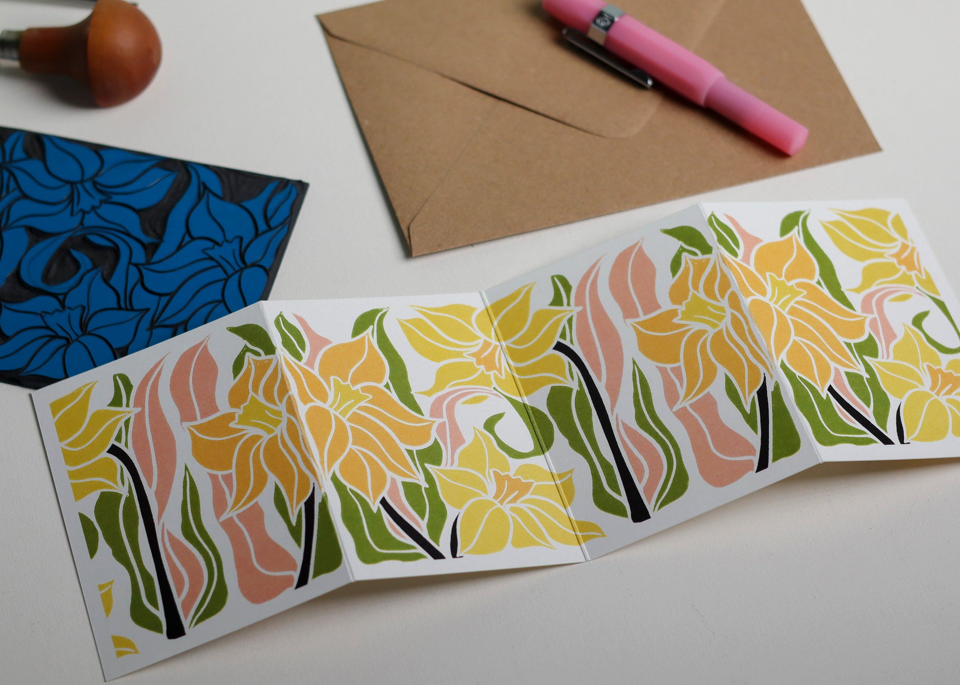 Hand-carved lino block shown beside finished Daffodil Garland card. Traditional linocut printmaking tools and original carved linoleum block displayed with a contemporary botanical portrait card. No AI, authentic craftsmanship from Norfolk studio