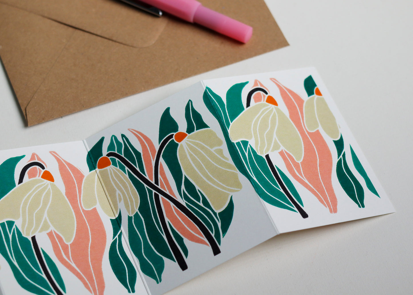 Snowdrop Garland card with C6 kraft envelope. Hand-carved botanical portrait greeting card in [pink, green and off-white on recycled cardstock, photographed with eco-friendly envelope, ready for posting