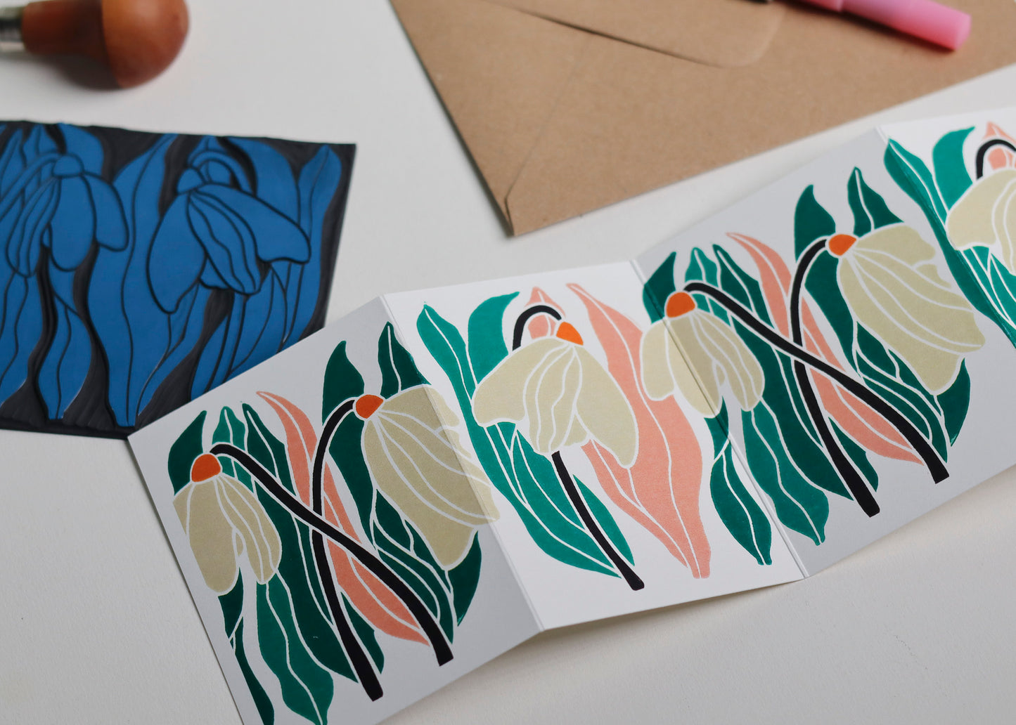 Hand-carved lino block shown beside finished snowdrop Garland card. Traditional linocut printmaking tools and original carved linoleum block displayed with contemporary pink and green botanical portrait card. No AI, authentic craftsmanship from Norfolk studio.