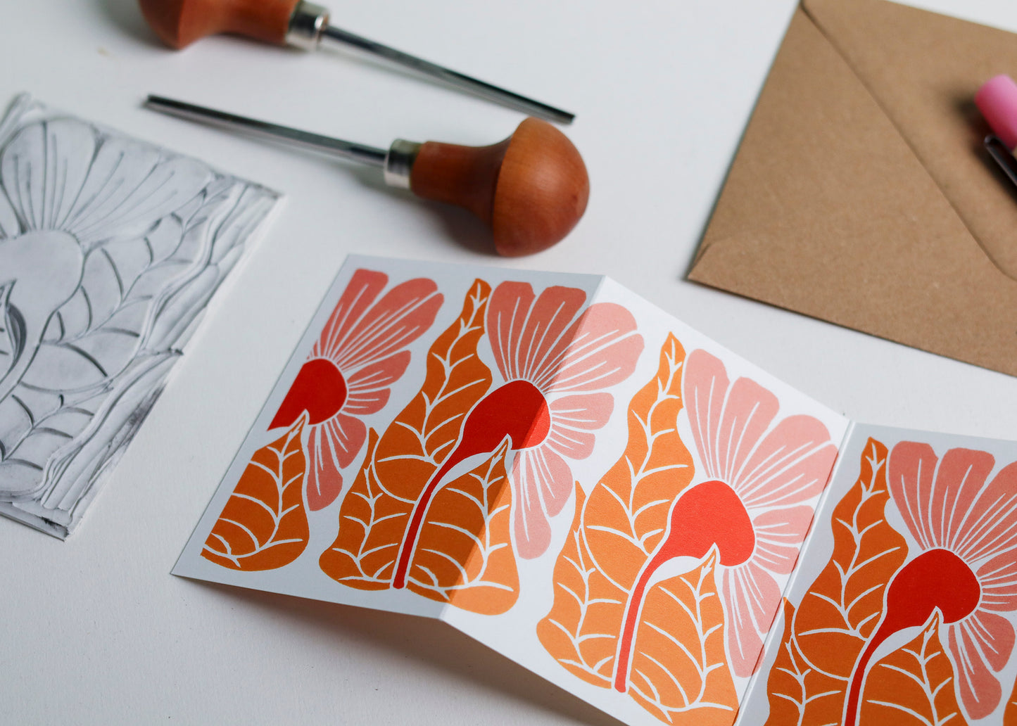 Hand-carved lino block shown beside finished Golden Autumn Garland card. Traditional linocut printmaking tools and original carved linoleum block displayed with contemporary coral pink colour botanical portrait card. No AI, authentic craftsmanship from Norfolk studio.