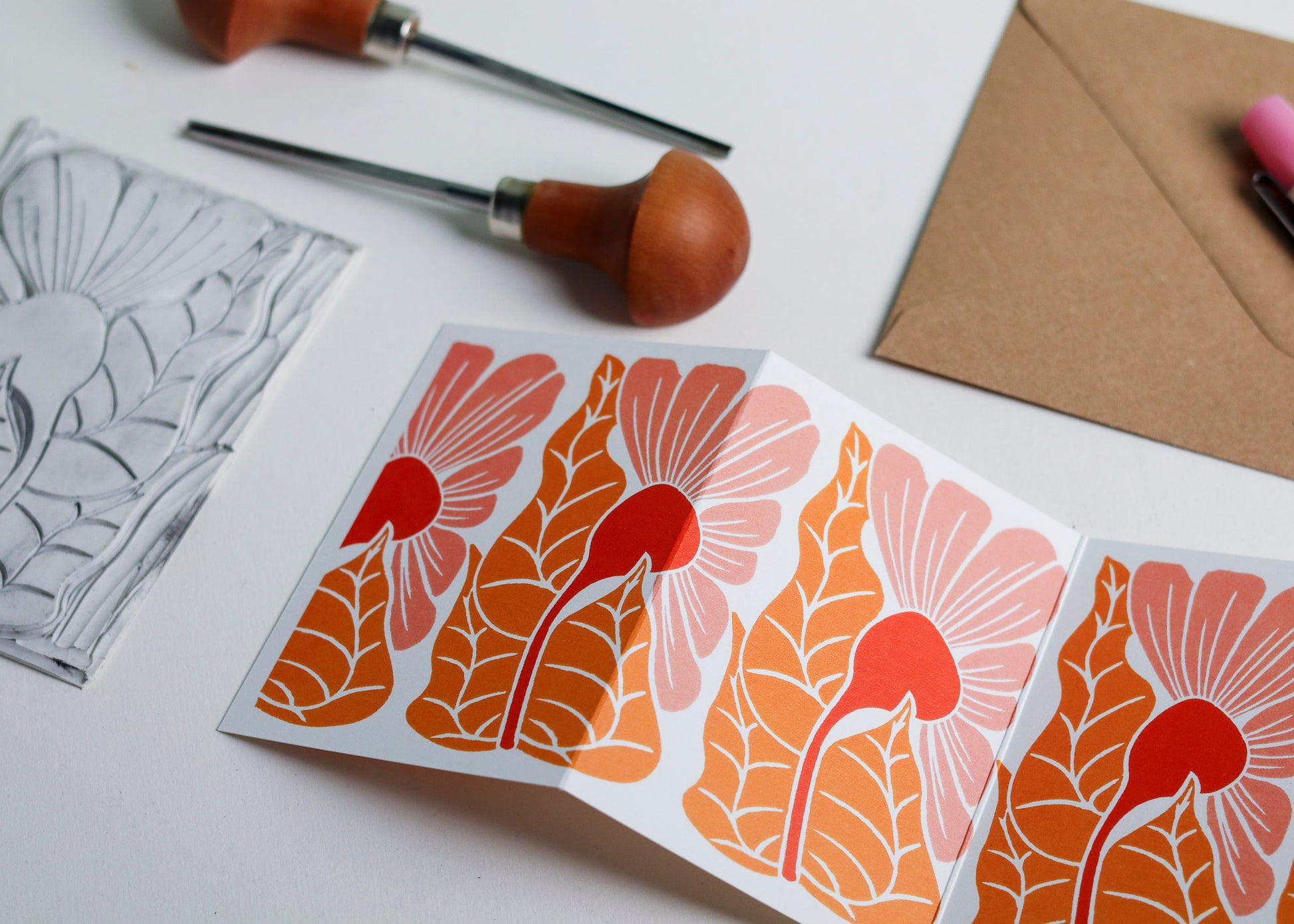 Hand-carved lino block shown beside finished Golden Autumn Garland card. Traditional linocut printmaking tools and original carved linoleum block displayed with contemporary coral pink colour botanical portrait card. No AI, authentic craftsmanship from Norfolk studio.