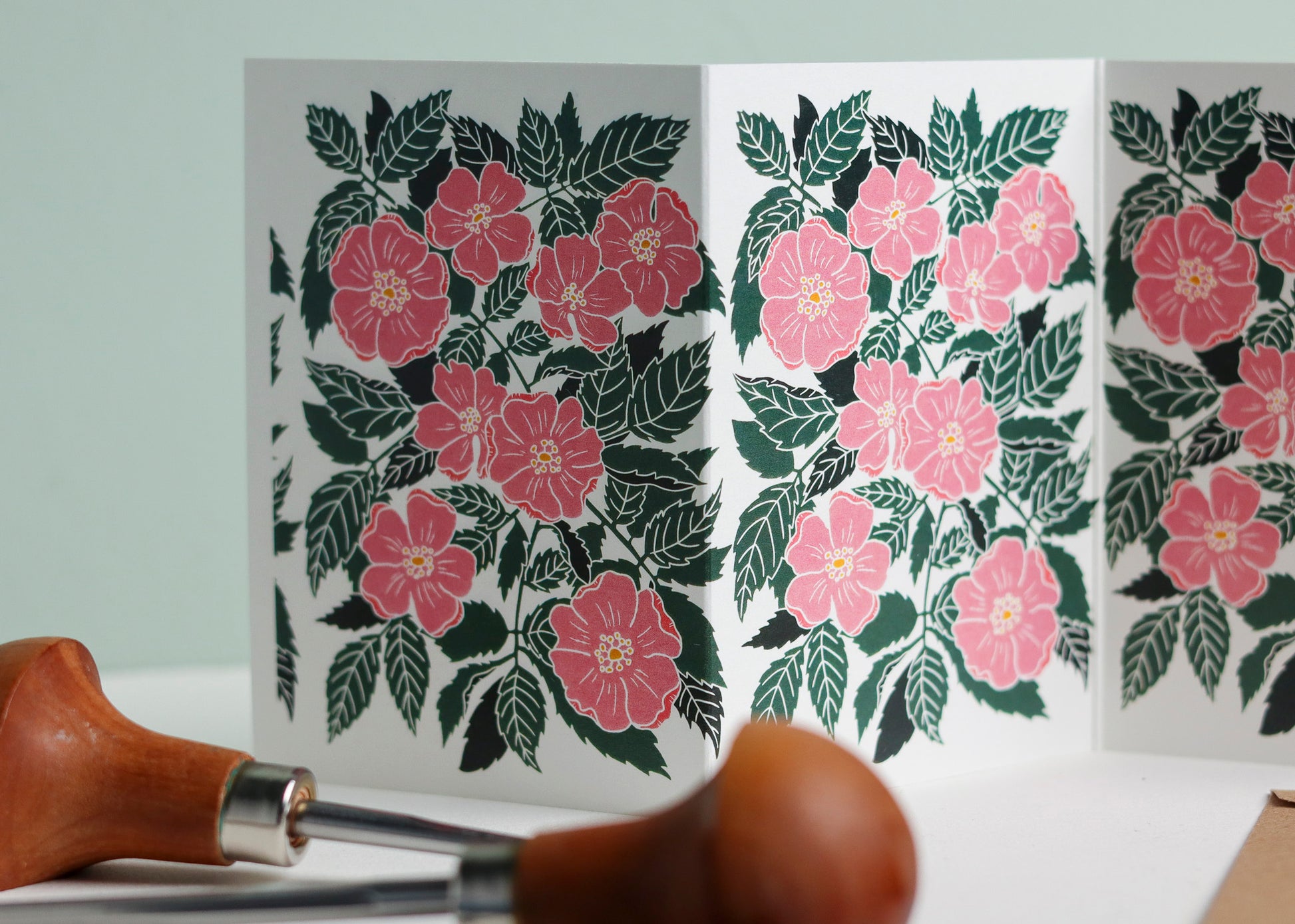 Close-up detail of hand-carved wild rose design on Garland card. Bold pink and green colours and graphic linocut linework showing botanical accuracy and contemporary colour palette