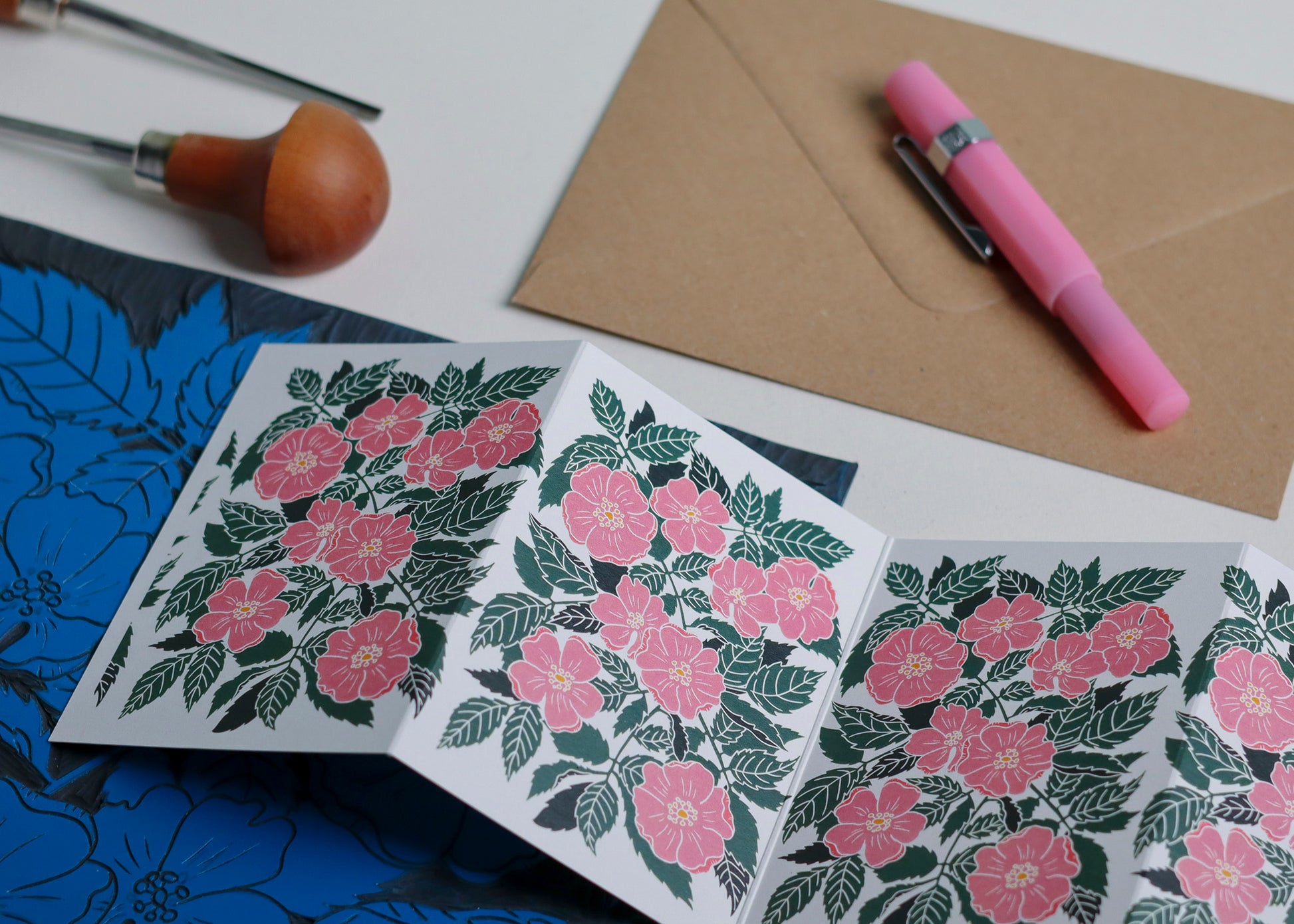 Dog Rose Garland card with C6 kraft envelope. Hand-carved botanical portrait greeting card in coral pink and forest green on recycled cardstock, photographed with eco-friendly envelope, ready for posting.