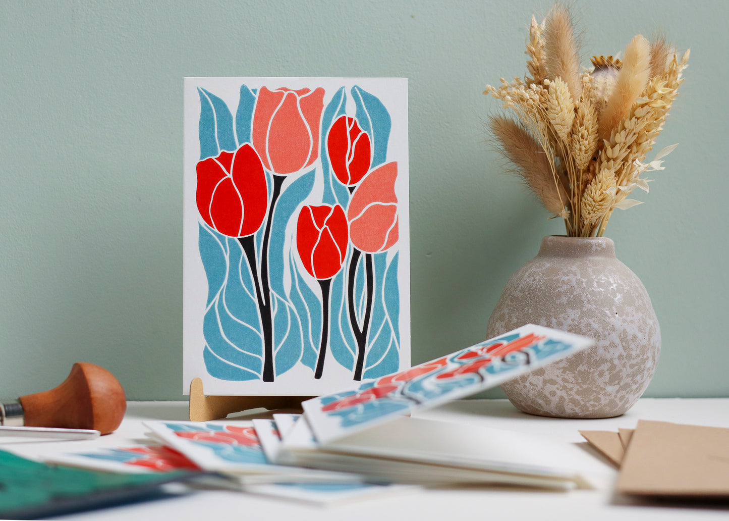 Card with red tulips design on a stand next to a vase with dried plants on a light surface.