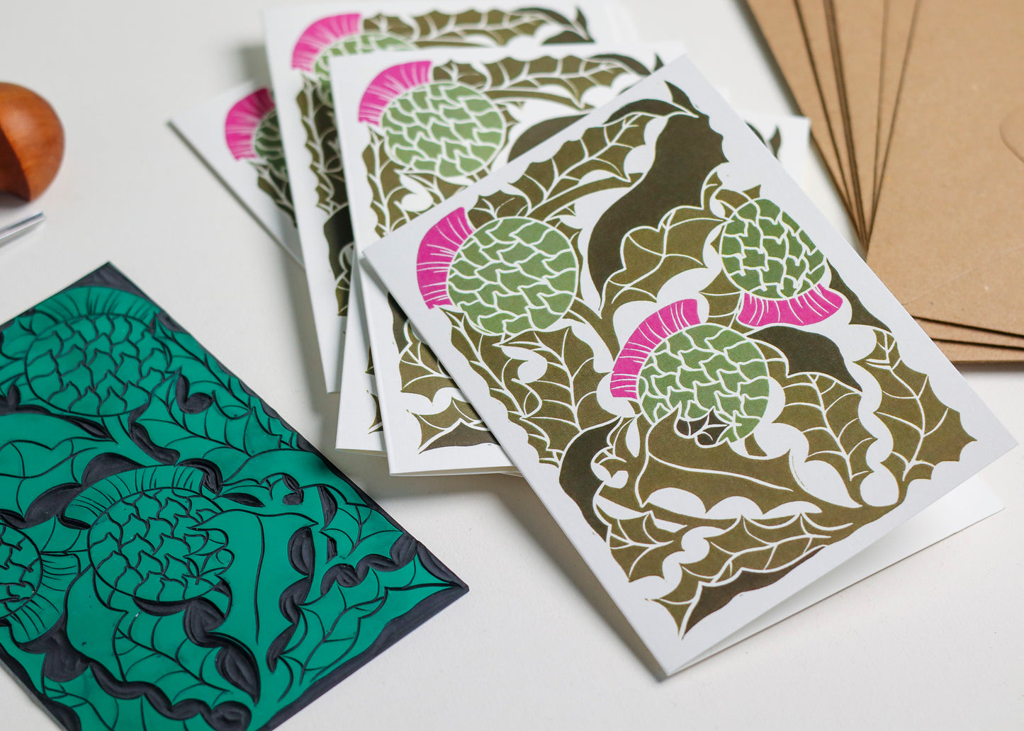 Hand-carved lino block displayed with finished Scottish Thistle notecard from set of 8. Traditional relief printmaking block showing carved botanical design alongside contemporary printed card. Authentic linocut process, Norfolk countryside studio.