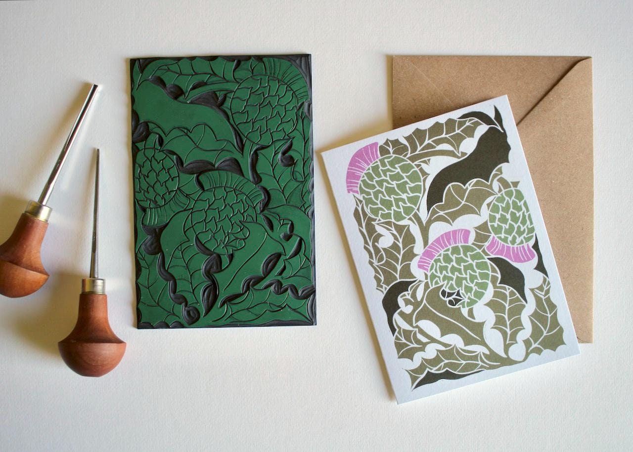 A linocut thistle flower and leaves design digitally printed onto luxury cotton card in a traditional green and purple colourway