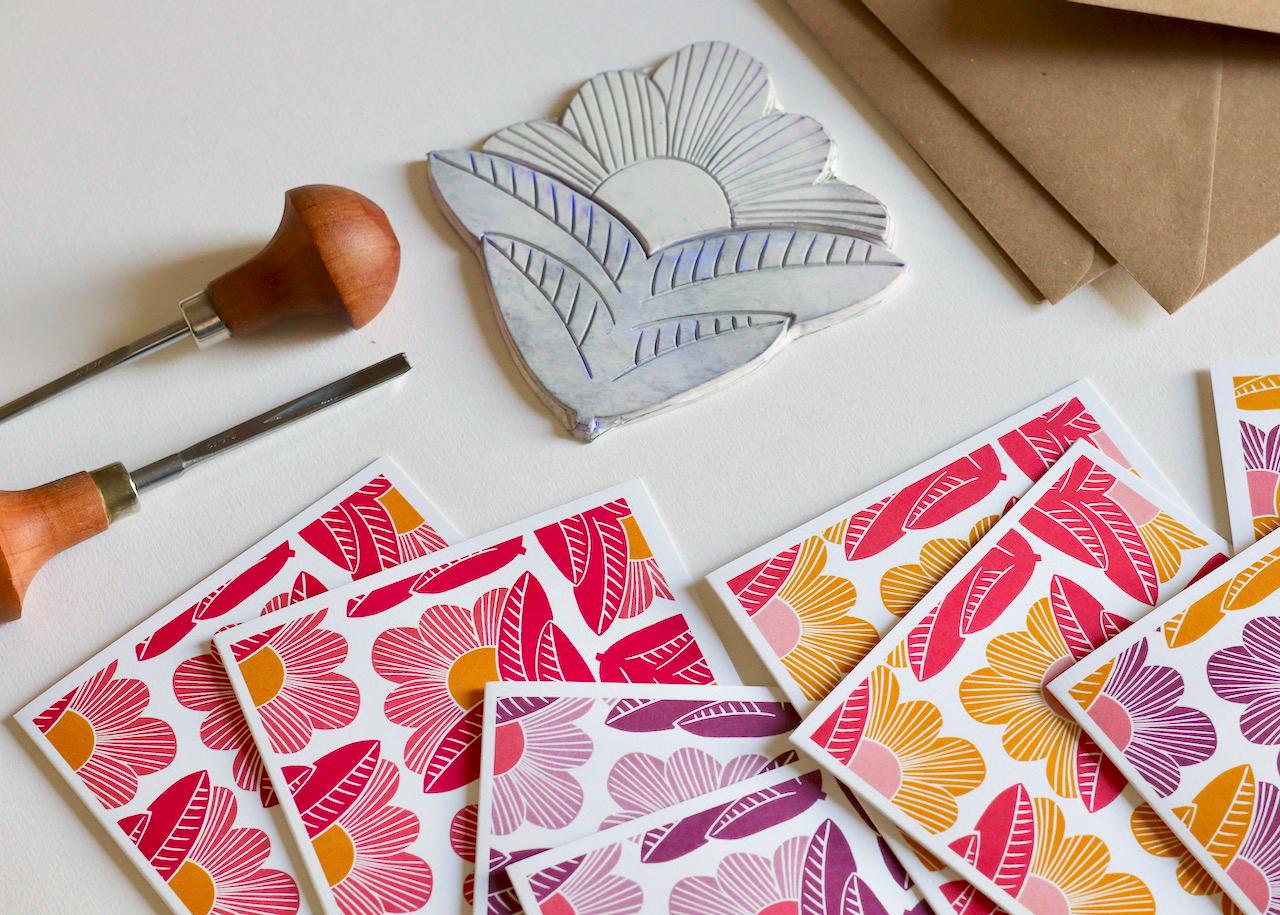 Small batch printed linocut notecard featuring abstract pink and golden yellow flowers with geometric petals and linear details on white cardstock, shown with kraft envelope