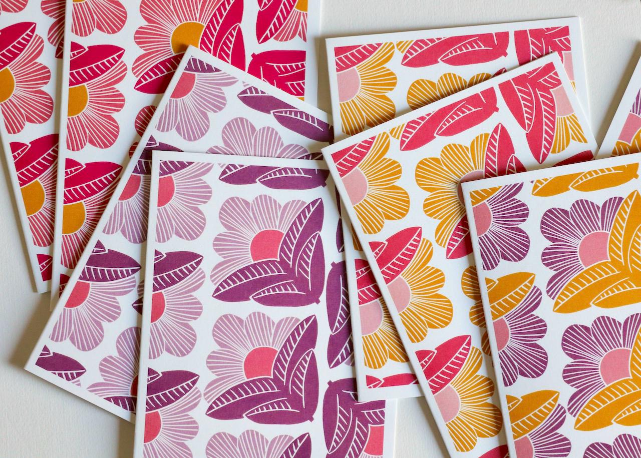 Flat lay of modern botanical notecard set displaying the &#39;Sunrise Floral&#39; pattern - a contemporary linocut design with stylized flowers in warm pink and yellow tones